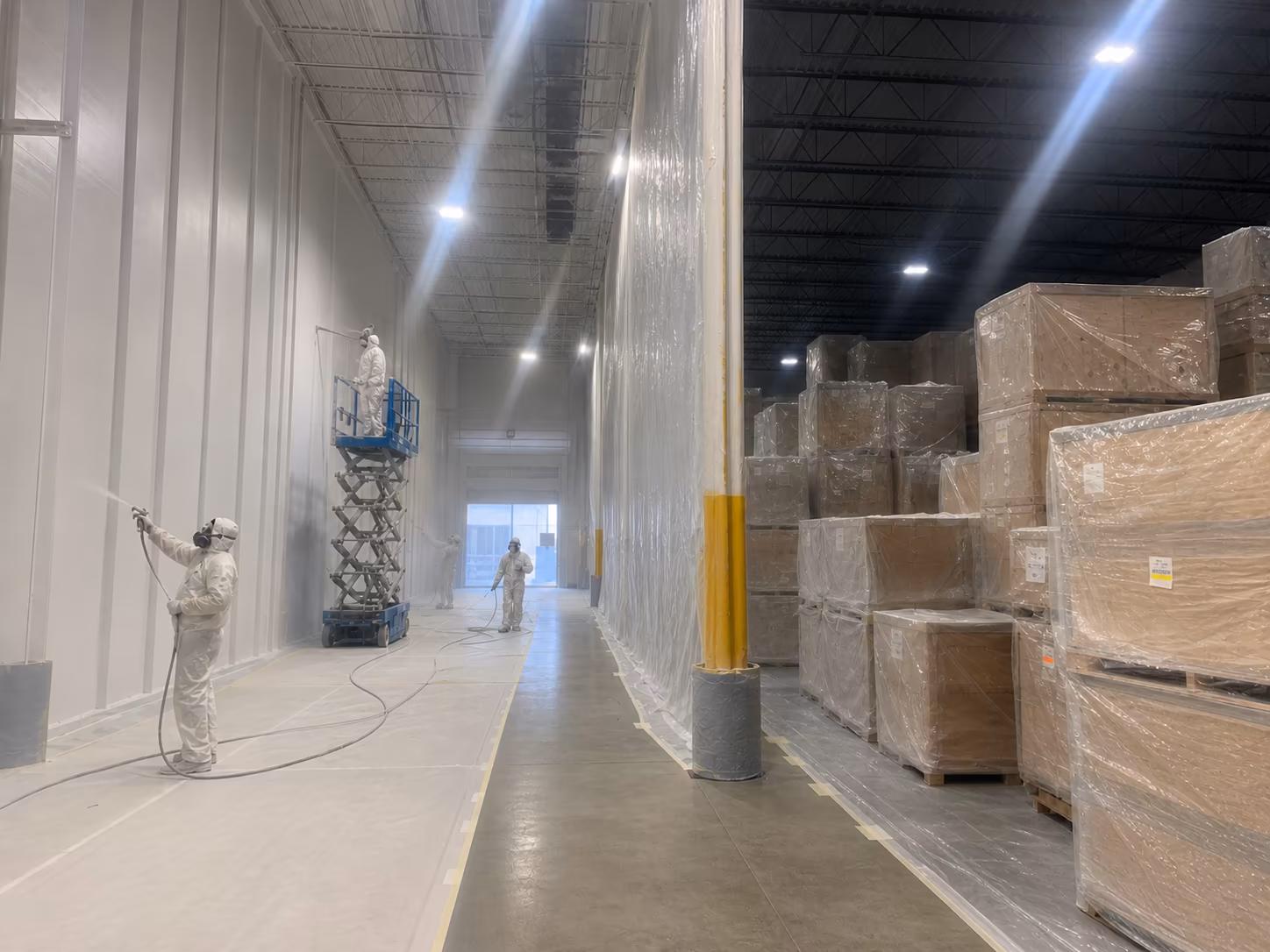 City Decor team in protective spray suits painting a large warehouse interior — scissor lift, masked-off pallets and dramatic warehouse lighting